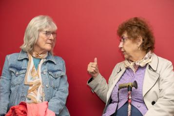 Two women talk to each other infront of a red wall.