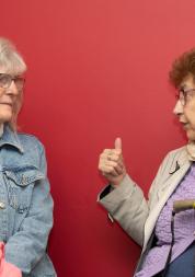 Two women talk to each other infront of a red wall.